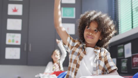 Diverse-happy-schoolchildren-sitting-at-desk-and-raising-hands-in-school-classroom