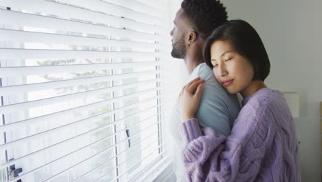 Happy-diverse-couple-embracing-and-looking-through-window-in-bedroom