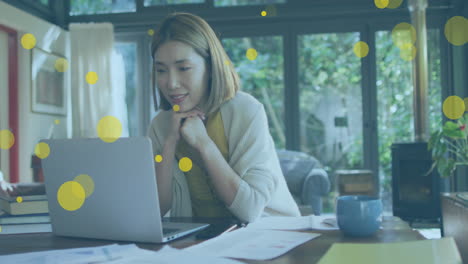 Animation-of-yellow-spots-floating-over-asian-woman-having-a-video-call-on-laptop-at-home