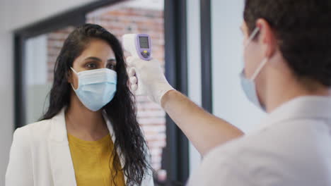 Man-wearing-face-mask-checking-temperature-of-woman-at-office
