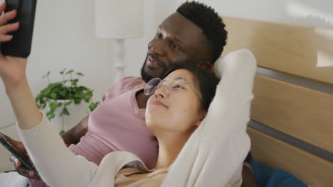 Happy-diverse-couple-using-smartphone-and-lying-in-bedroom