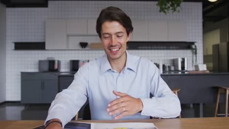 Smiling-caucasian-businessman-having-video-chat-going-through-paperwork-in-workplce-kitchen