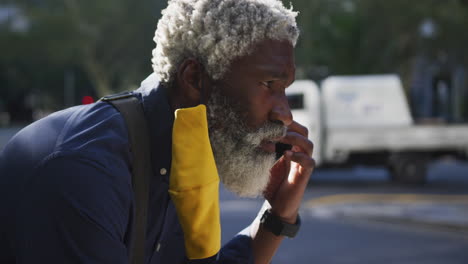 Close-up-of-african-american-senior-man-wearing-face-mask-talking-on-smartphone-on-the-road