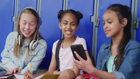 Video-of-three-happy,-diverse-schoolgirls-looking-at-smartphone,-sitting-by-lockers,-copy-space