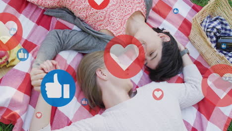 Animation-of-hearts-and-like-icons-over-caucasian-female-couple-having-picnic