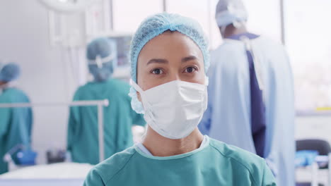 Video-portrait-of-biracial-female-surgeon-in-face-mask-in-operating-theatre,-with-copy-space