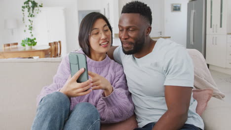 Diverse-couple-sitting-on-couch-and-using-smartphone-in-living-room