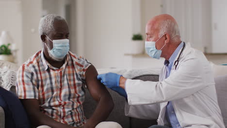 Senior-caucasian-male-doctor-vaccinating-senior-african-american-man-at-home-both-wearing-face-masks