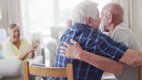 Two-happy-diverse-senior-men-hugging-in-living-room,-with-diverse-senior-friends,-slow-motion