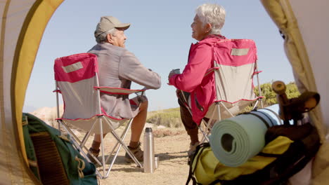 Happy-senior-biracial-couple-sitting-at-tent-in-mountains-and-kissing,-in-slow-motion