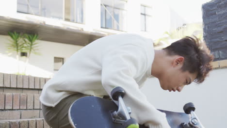 Asian-male-teenager-sitting-with-skateboard-on-sunny-day-in-garden