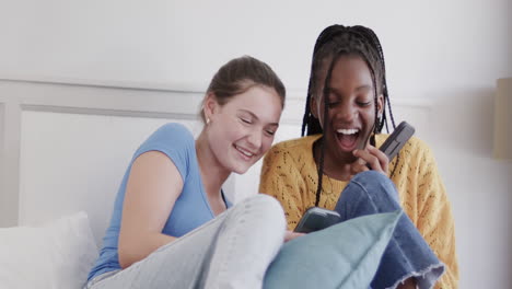 Happy-diverse-teenage-female-friends-lying-on-bed-using-smartphones-and-laughing