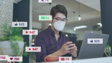 Animation-of-social-media-icons-over-asian-man-wearing-face-mask-using-smartphone-at-office