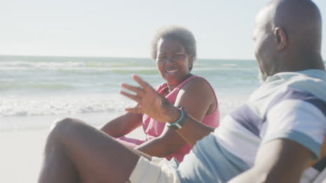 Happy-senior-african-american-couple-sitting-on-deck-chairs-and-talking-at-beach,-in-slow-motion