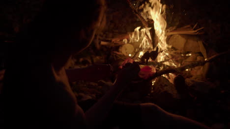 Slow-motion-silhouette-of-man-holding-skewer-of-food-over-open-campfire-to-cook-it