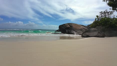 Slow-motion-of-waves-crashing-on-white-sandy-beach-and-granite-stones-on-a-sunny-day,-Mahe-Seychelles