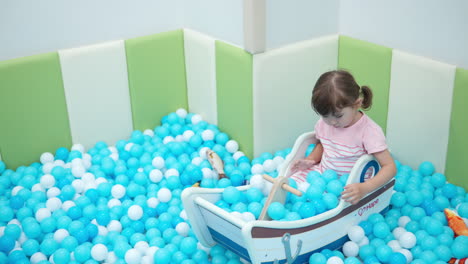 Little-girl-plays-in-a-dry-pool-with-white-and-blue-balls,-sitting-inside-wooden-toy-boat