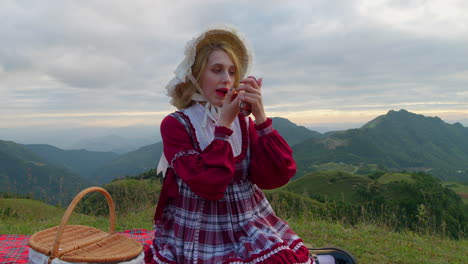 female-blonde-model-in-traditional-old-fashioned-style-clothing-doing-make-up-with-lipstick-and-portable-mirror-in-stunning-outdoor-mountains-natural-location