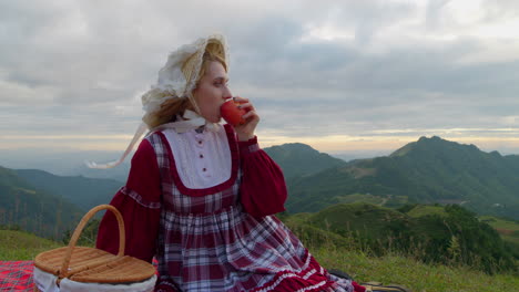 young-blonde-female-model-in-traditional-old-fashioned-renaissance-clothing-dress-eating-a-red-apple-in-slow-motion-while-sitting-alone-in-mountains-landscape