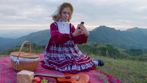 female-blonde-model-in-old-fashioned-traditional-clothing-holding-a-modern-smartphone-taking-a-selfie-in-nature-while-having-lunch-alone