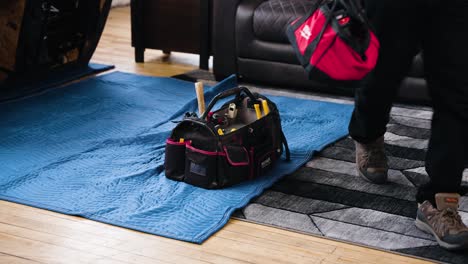 A-Male-Handy-Man-Furniture-Repair-Trades-Carpenter-Leaves-his-Tool-Bag-Full-of-Equipment-Hammer-Plyer-Drill-Behind-in-a-Home-Living-Room-on-an-Industrial-Padded-Blanket