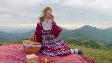 young-blonde-female-model-in-traditional-old-fashioned-renaissance-dress-clothing-sitting-alone-having-lunch-and-eating-a-red-apple-in-mountains-landscape-historical-reproduction