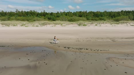 Aerial-view-with-a-young-longhaired-girl-riding-a-bike-on-the-sandy-beach,-sunny-day,-white-sand-beach,-active-lifestyle-concept,-wide-drone-dolly-shot-moving-right,-slow-zoom-in