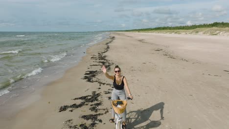 Aerial-view-with-a-young-longhaired-girl-riding-a-bike-on-the-sandy-beach,-waving-hand,-sunny-day,-white-sand-beach,-active-lifestyle-concept,-wide-drone-shot-moving-backward