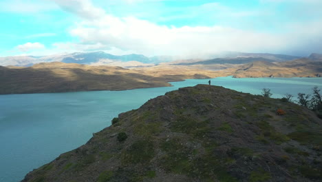 Aerial-drone-view-of-a-hiker-standing-on-top-of-a-mountain-over-looking-a-scenic-lake-in-Patagonia