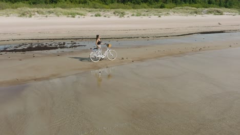 Aerial-view-with-a-young-longhaired-girl-standing-with-a-bike-on-the-sandy-beach,-sunny-day,-white-sand-beach,-active-lifestyle-concept,-wide-ascending-drone-shot-moving-backward