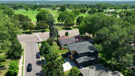 An-aerial-view-of-a-suburban-neighborhood-with-a-house-equipped-with-solar-panels,-a-crosswalk-leading-to-a-park-and-golf-course-area,-and-a-car-driving-on-the-road