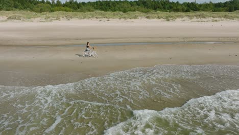 Aerial-view-with-a-young-longhaired-girl-riding-a-bike-on-the-sandy-beach,-sunny-day,-white-sand-beach,-active-lifestyle-concept,-wide-drone-dolly-shot-moving-right