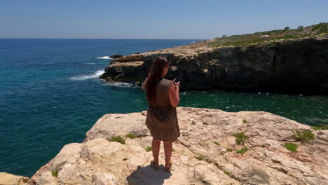 Frau-Steht-An-Der-Klippe-Und-Hält-Ihr-Mobiltelefon-An-Der-Westküste-Von-Malta,-Spanien