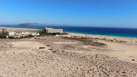 Aerial-view-traveling-in-to-the-beach-and-hotel-of-Corralejo-in-Fuerteventura,-seeing-the-road-that-crosses-it