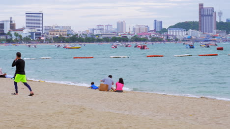 Tourists-by-the-beach-in-Pattaya,-facing-the-Gulf-of-Thailand-littered-with-speed-boats,-jetskis,-and-yatchs-for-water-sports,-and-the-business-center-of-Chonburi-province-in-the-background,-Thailand