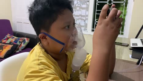 Un-Niño-Asiático-Usa-Un-Nebulizador-Inhalador-Mientras-Juega-Con-Su-Teléfono-Inteligente.
