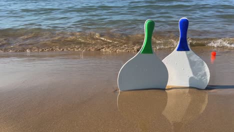 Beach-Tennisschläger-Und-Rote-Bälle-Am-Sandstrand-Mit-Wellen,-Die-Sich-Am-Ufer-Brechen