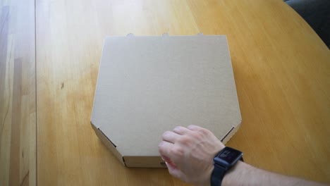 the-handheld-shot-of-man's-hand-opening-delivered-Italian-pizza-in-a-cardboard-box-on-the-table-at-home