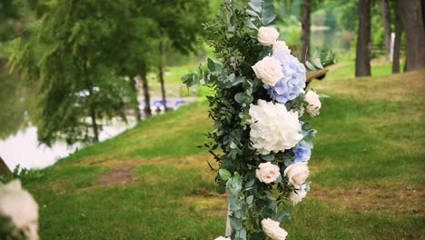 revealing-shot-of-blue-and-white-wedding-ceremony-flowers-on-a-metal-stand-with-green-leaves-in-the-nature-near-the-lake