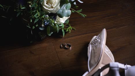Top-view-of-the-wedding-rings,-bridal-shoes-and-flower-bouquet-on-a-dark-wooden-surface,-rotating-shot