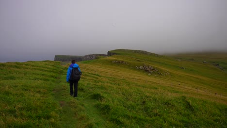 Static-rear-shot-of-man-hiking-through-green-meadow-towards-foggy-cliffs,-Mykines