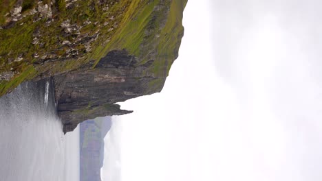 Tilt-down-shot-of-Trollkonufingur-emerging-from-ocean-next-to-cliffs-at-Vagar,-Faroe-Islands