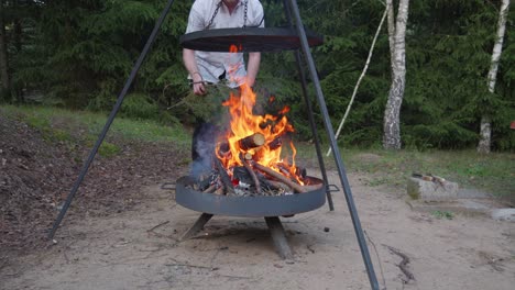 Man-in-white-shirt-Putting-Some-Woods-into-campfire-grill-or-Fire-pit-grill-with-burning-wood-in-the-firebox
