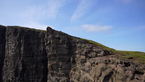 Man-in-yellow-raincoat-climbs-Traelanipa-cliff-on-sunny-day,-Faroe-Islands