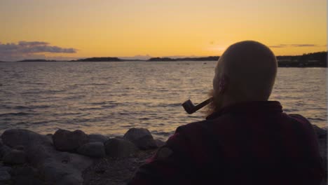 A-man-is-sitting-on-a-rocky-beach-looking-out-at-the-ocean-with-pipe-in-his-mouth-the-sky-is-golden-sunset-which-casts-an-orange-hue-across-the-horizon-creating-a-silhouette-of-the-person-calm-smoking
