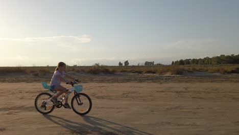 Little-girl-riding-a-bicycle-on-a-sandy-road-as-the-sun-shines-at-dusk,-Catalonia,-Spain