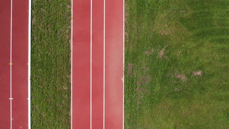 Drone-top-down-pan-across-empty-red-rubber-of-track-lined-with-mowed-grassy-field