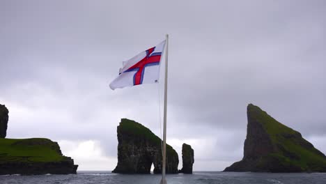 Die-Färöische-Flagge-Flattert-Im-Wind-Mit-Den-Felsnadeln-Von-Drangarnir-Und-Der-Insel-Tindholmur-Im-Hintergrund
