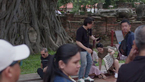 Fesselnde-Szene-Mit-Menschen,-Die-Aufmerksam-Einer-Historischen-Erzählung-Vor-Dem-Kopf-Des-Buddha-Lauschen,-Der-In-Den-Wurzeln-Eines-Baumes-In-Ayutthaya,-Thailand,-Eingebettet-Ist