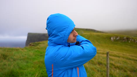 Medium-view-of-man-covering-head-with-blue-jacket-hood,-walking-along-Mykines-coastline